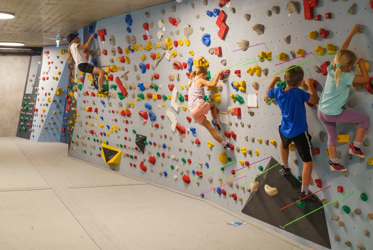 Eine Gruppe von Kindern im Alter von 5 bis 10 Jahren klettert gleichzeitig an verschiedenen Routen in einer Boulderhalle. Alle Kinder sind an der Wand, während bunte Kletterwände eine lebhafte Atmosphäre schaffen.